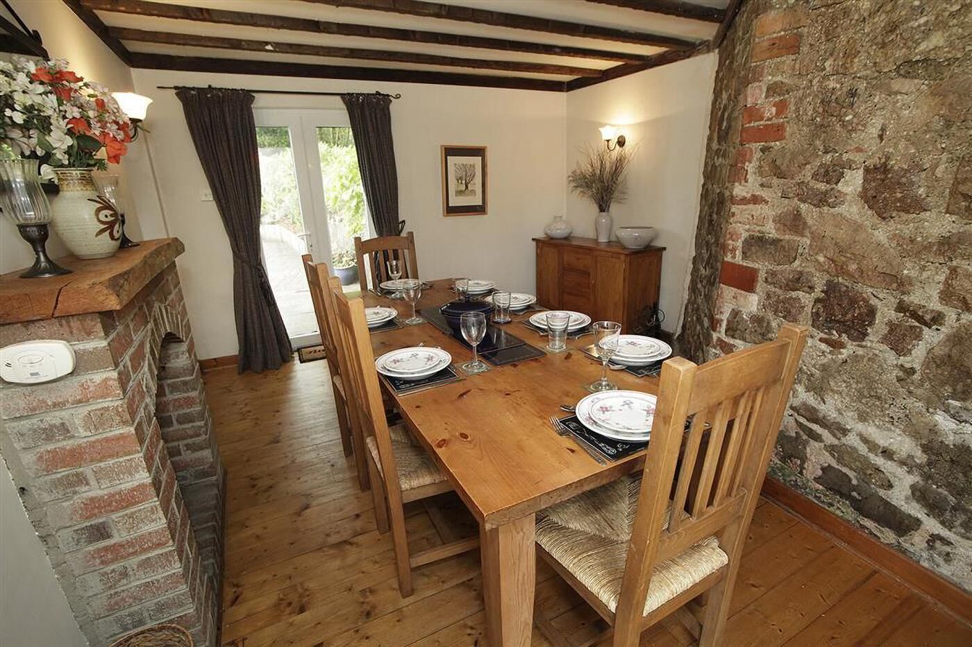 Une salle à manger avec une table en bois dressée pour quatre personnes, entourée de chaises, d’une cheminée en briques et d’un mur en pierre.