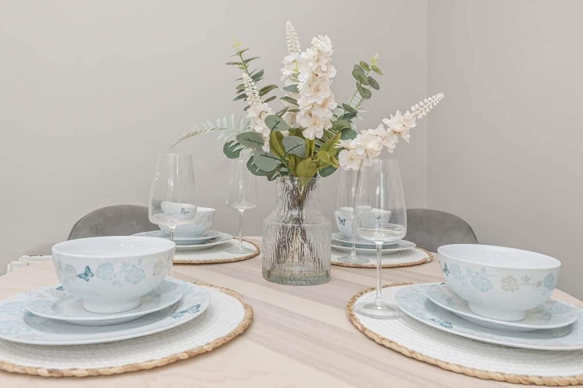Et spisebord dækket med skåle og tallerkener med et hvidt blomstermønster, en klar glasvase med hvide blomster og vinglas.