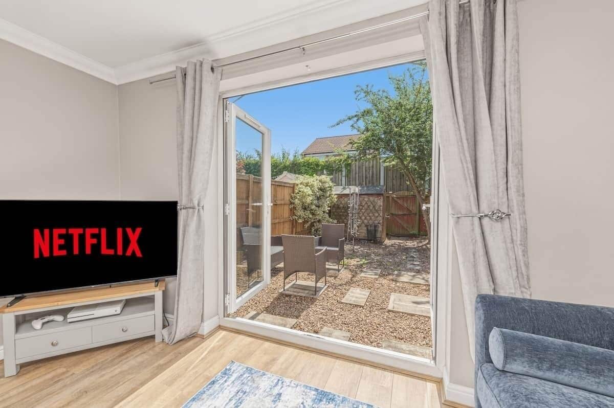 Et værelse med et fjernsyn, der viser 'NETFLIX', en skydedør til et udendørs område med terrasse, og en grå sofa.