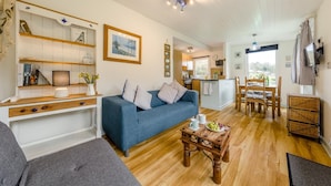A living room with a blue sofa, wooden coffee table, and a dining area with chairs and a table. - 1 Bed in Caswell (oc-hh372) (Newton)