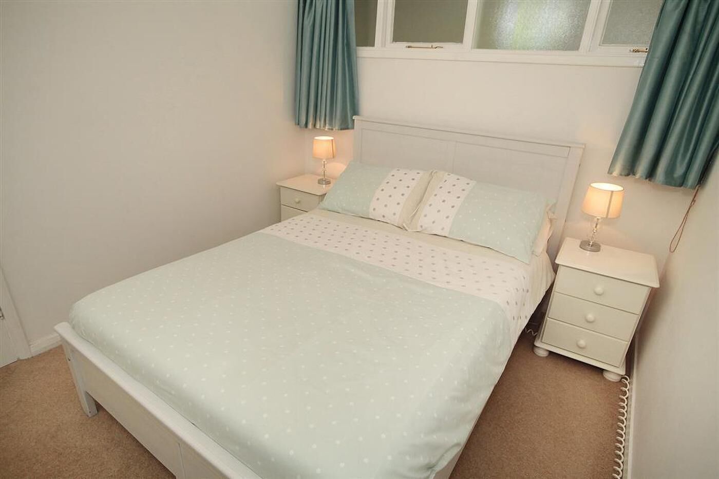 A single-bed room with a white headboard, two bedside tables with lamps, a window with curtains, and a carpeted floor.