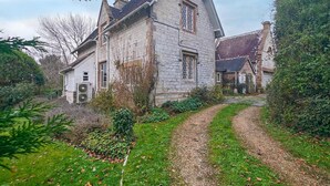 Een stenen huis met een steil dak, een grindpad dat naar de ingang leidt en een tuin met struiken en een kleine boom.