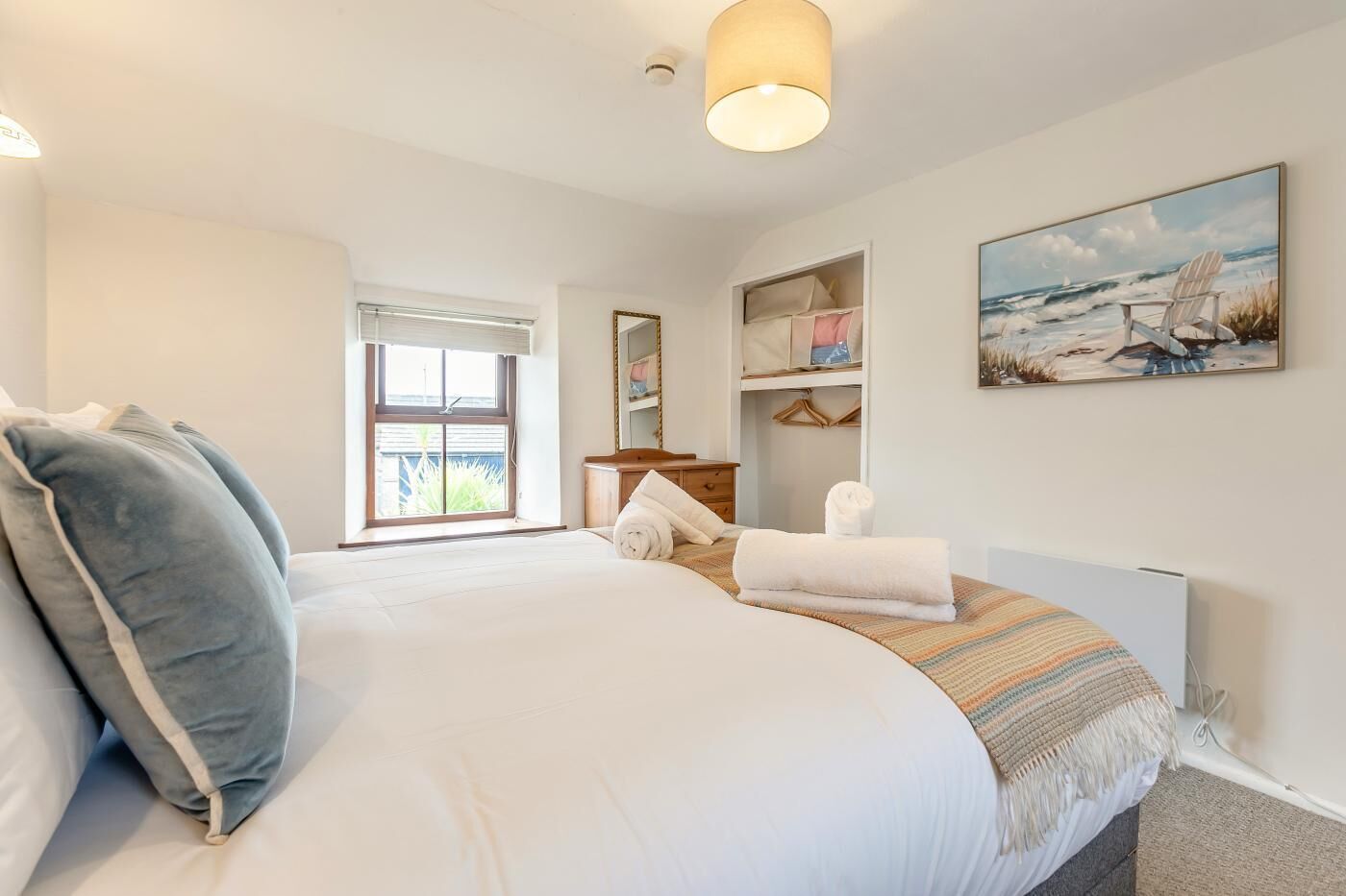 Een slaapkamer met een groot bed, een raam, een nachtkastje, een spiegel, een plank met handdoeken en een schilderij van een strandgezicht.