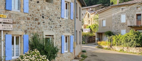 A stone building with blue shutters, a gravel path, and a sign indicating 'Juvinas' and 'Moulin-Lacoste'.
