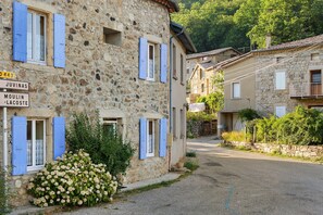 A stone building with blue shutters, a gravel path, and a sign indicating 'Juvinas' and 'Moulin-Lacoste'.