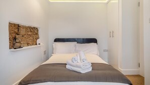 A neatly made bed with a brown headboard, white pillows, and a folded towel on the bed. There is a wall-mounted shelf with a stone-like decoration and a small lamp. - 4 Bed in Braunton (oc-n34220) (Braunton)