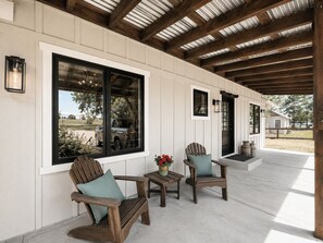 A covered porch with wooden chairs and a table, large windows, and a door. - The Grain House @ What The Farm (Loveland)