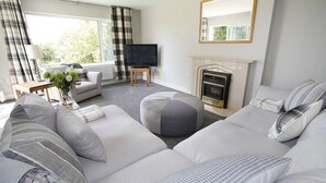 Una sala de estar moderna con un conjunto de sofá, un televisor de pantalla plana, una chimenea y un amplio ventanal con cortinas.
