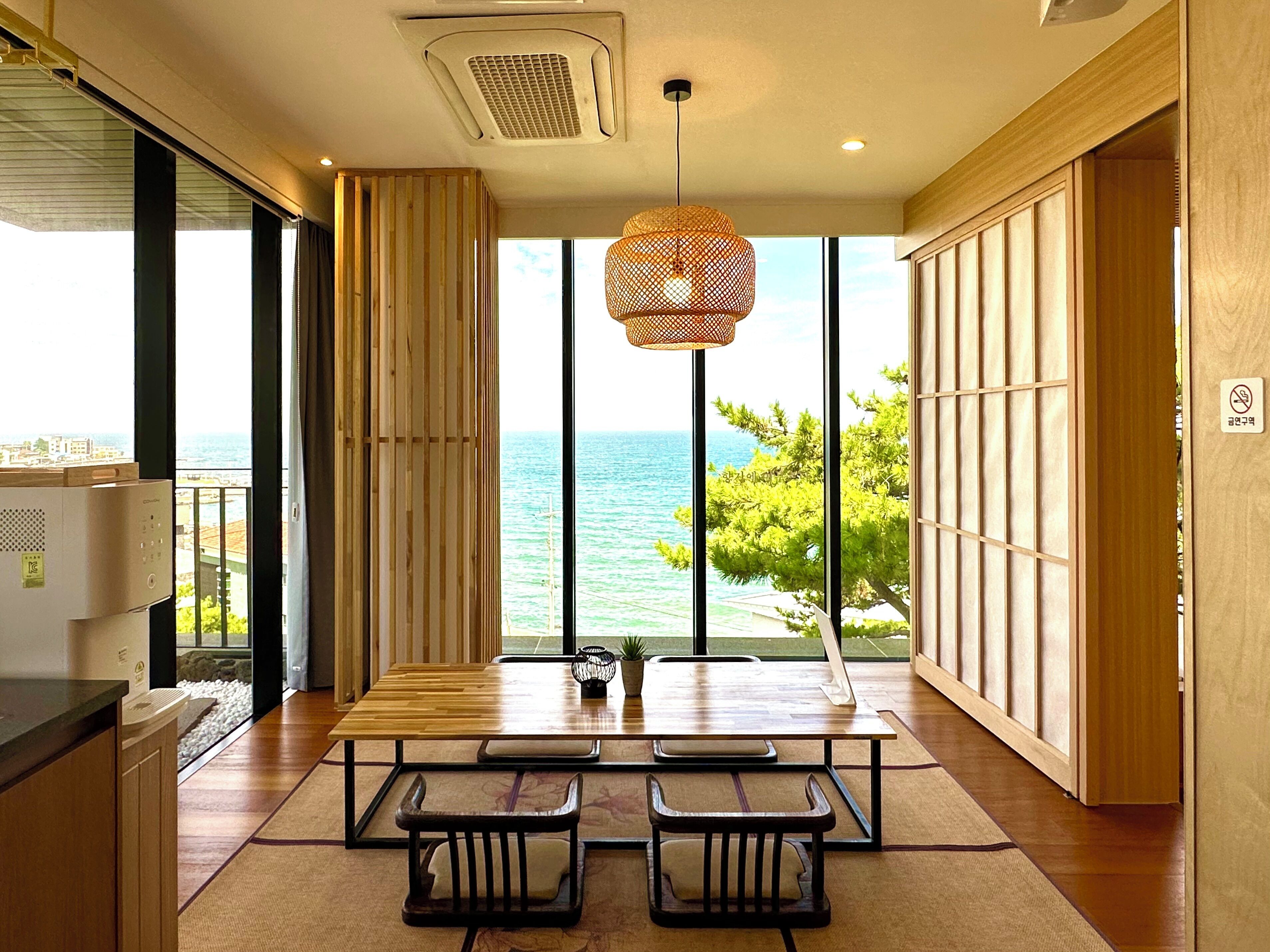 A modern kitchen with a large window overlooking the sea, wooden furniture, and a hanging light fixture.