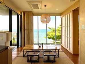 A modern kitchen with a large window overlooking the sea, wooden furniture, and a hanging light fixture.