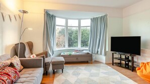 A living room with a sofa, ottoman, a flat-screen TV, a floor lamp, and a window with curtains. - 3 Bed in Ilfracombe (oc-s34501) (Ilfracombe)