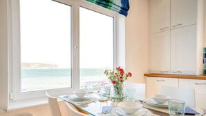 A dining area with a view of the sea, a table set with plates and glasses, a vase with flowers, and a kitchen with white cabinets. - 2 Bed in Swanage (oc-t34732) (Swanage)