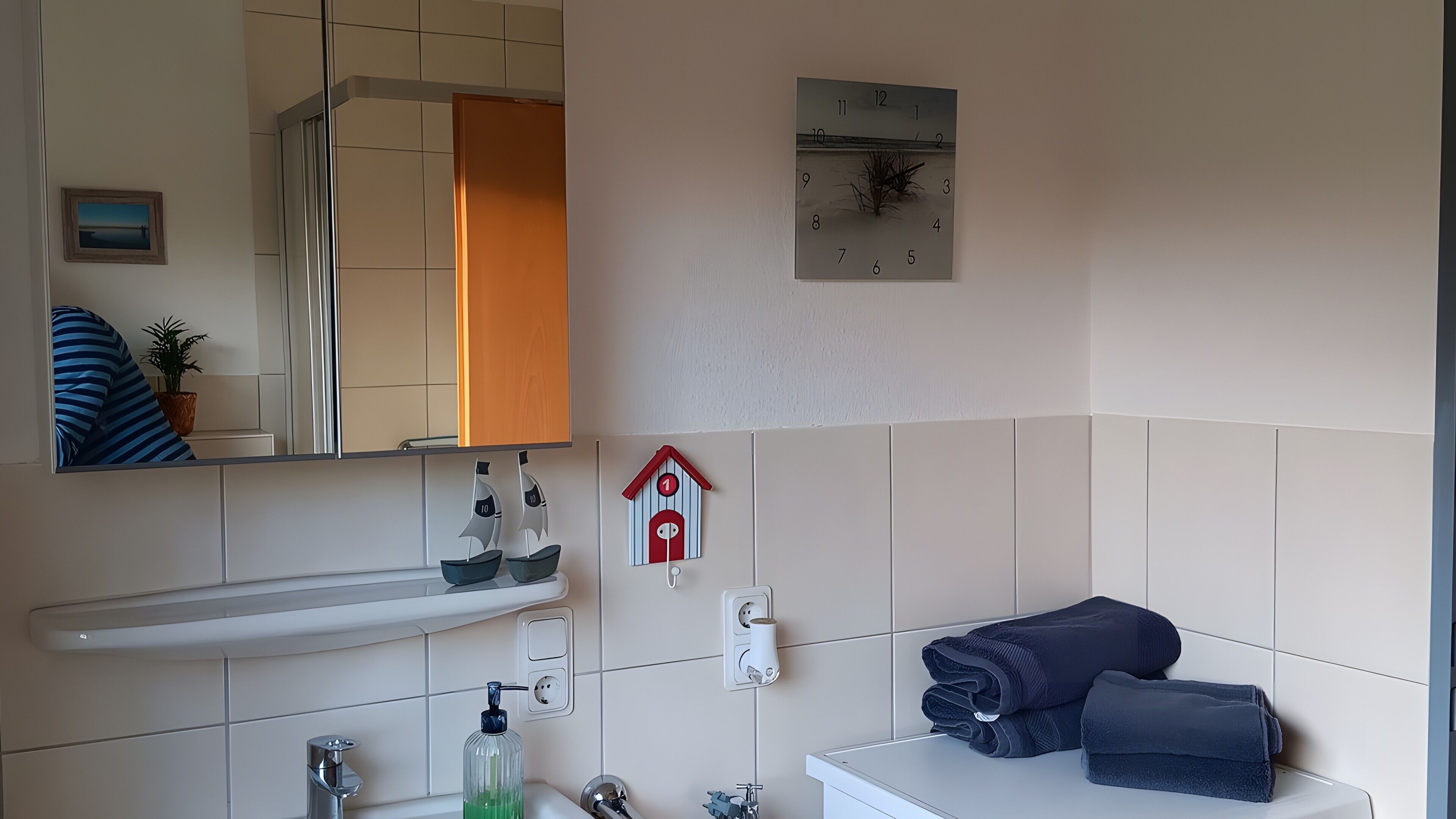 Ein Badezimmer mit Spiegel, einer Uhr, einem Handtuchhalter, einem Seifenspender und einer kleinen Topfpflanze.