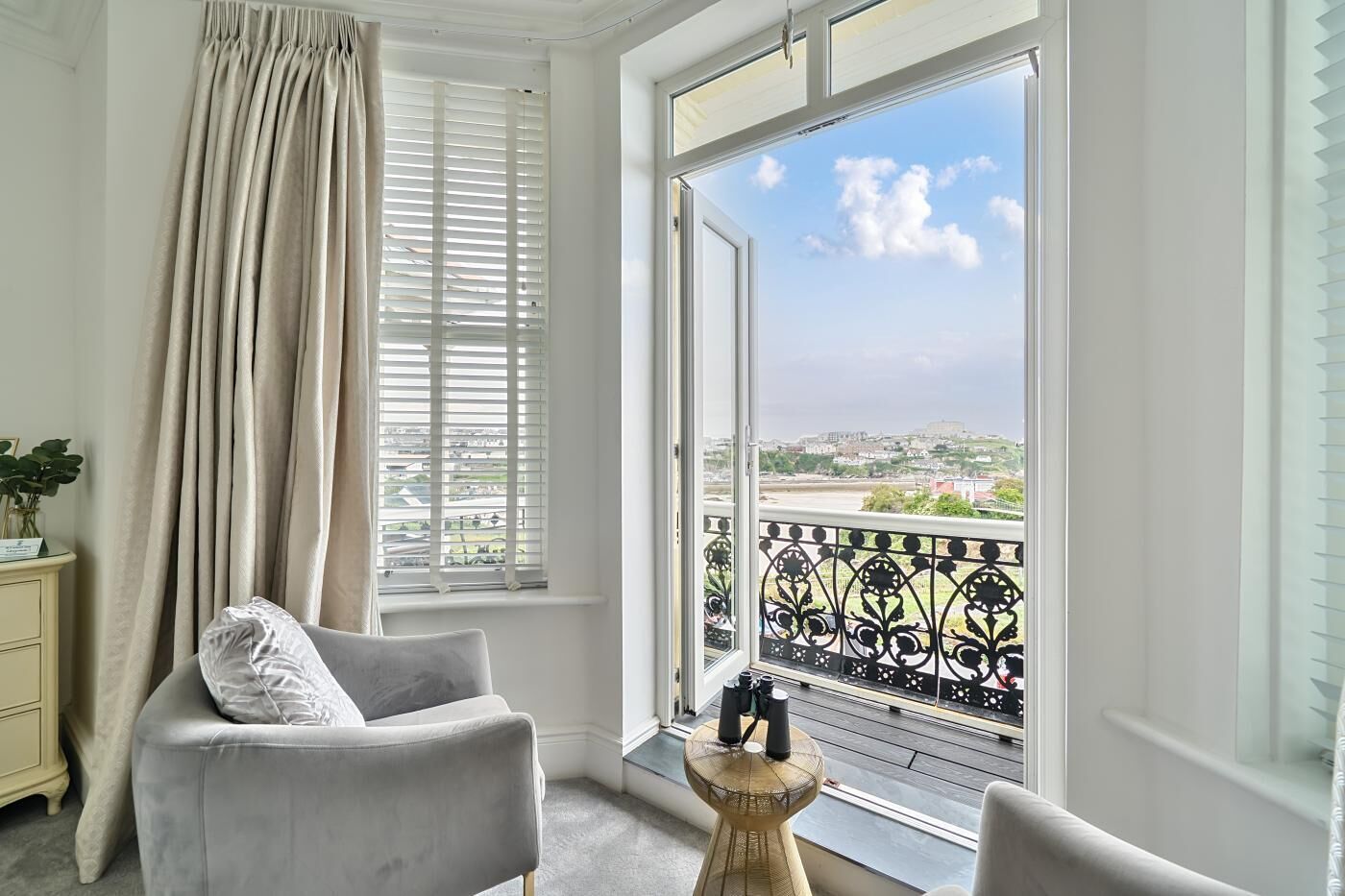 Ein Zimmer mit Balkon, einem Sessel, einem kleinen Tisch und Blick auf eine Stadtlandschaft.