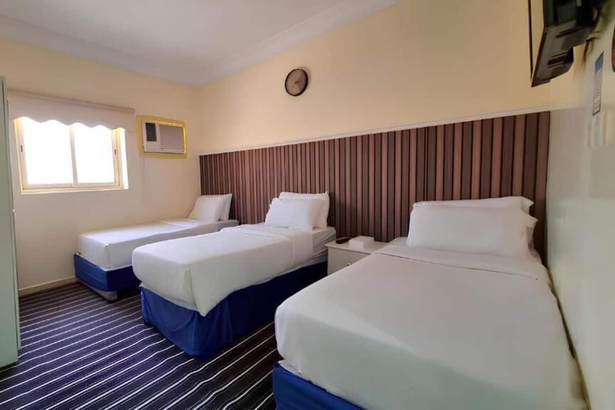 Quarto de hotel com duas camas, um guarda-roupa, uma janela com persianas, um relógio e uma televisão.
