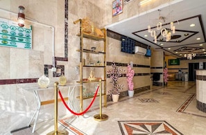 Um hall de hotel com piso de mármore, balcão de recepção, prateleira de exibição com moldura dourada e uma barreira de corda vermelha.
