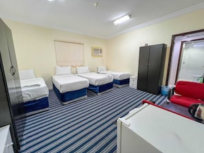 Quarto de hotel com quatro camas, uma escrivaninha, uma cadeira vermelha, um guarda-roupa e uma janela com persianas.
