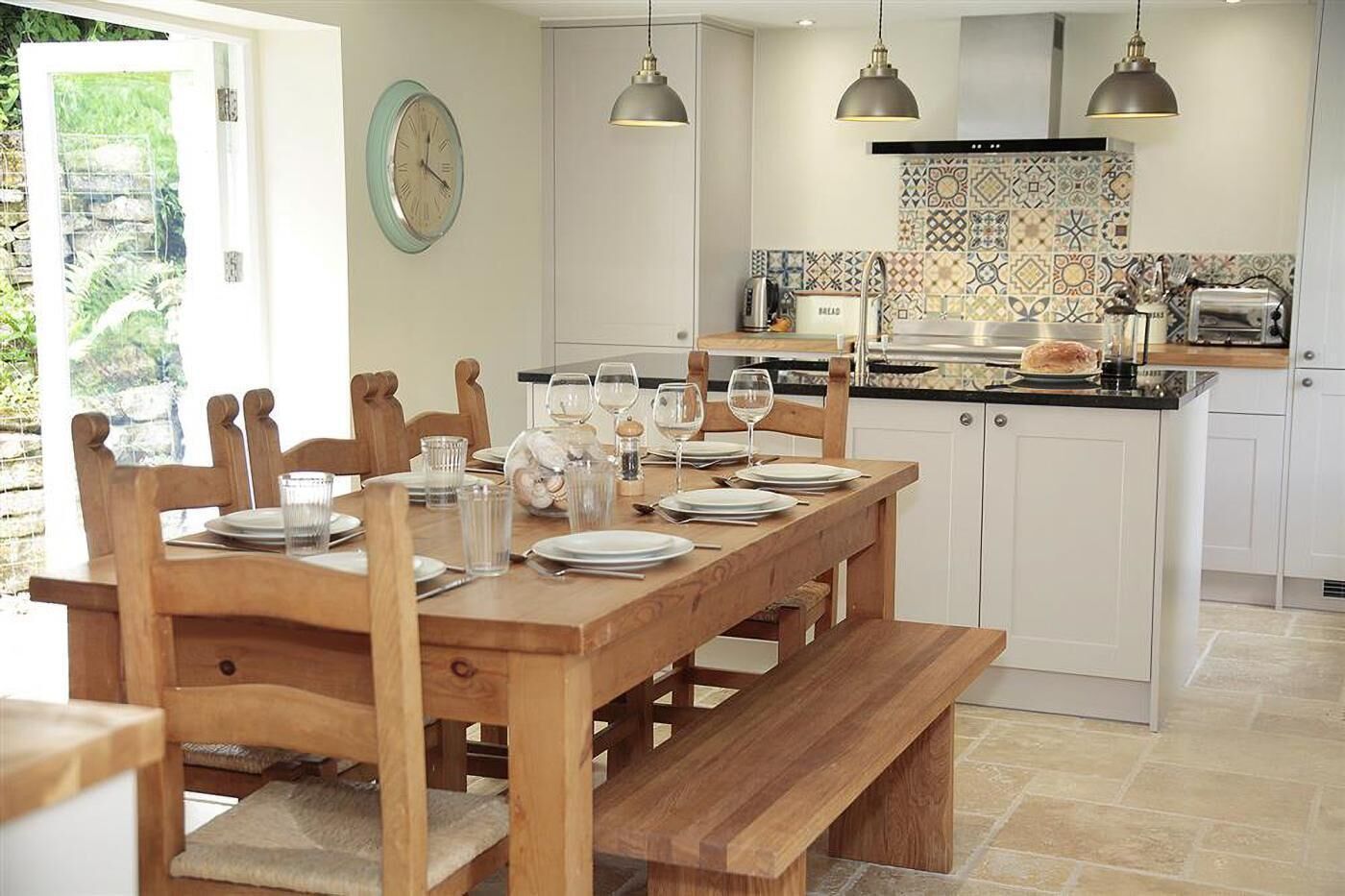 Un coin repas avec une table en bois dressée pour six personnes, une cuisine avec un mur recouvert de carreaux et une horloge au mur.