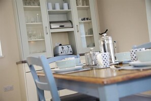 A dining area with a table set for two, a cabinet with glassware, and a kettle on the counter. - 4 Bed in Gower (oc-hh45) (Pilton Rhossili)