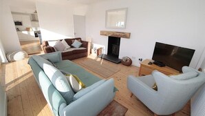 Ein modernes Wohnzimmer mit einer Couch, einem Sessel und einem Fernseher, einem Kamin sowie einer Küche im Hintergrund.