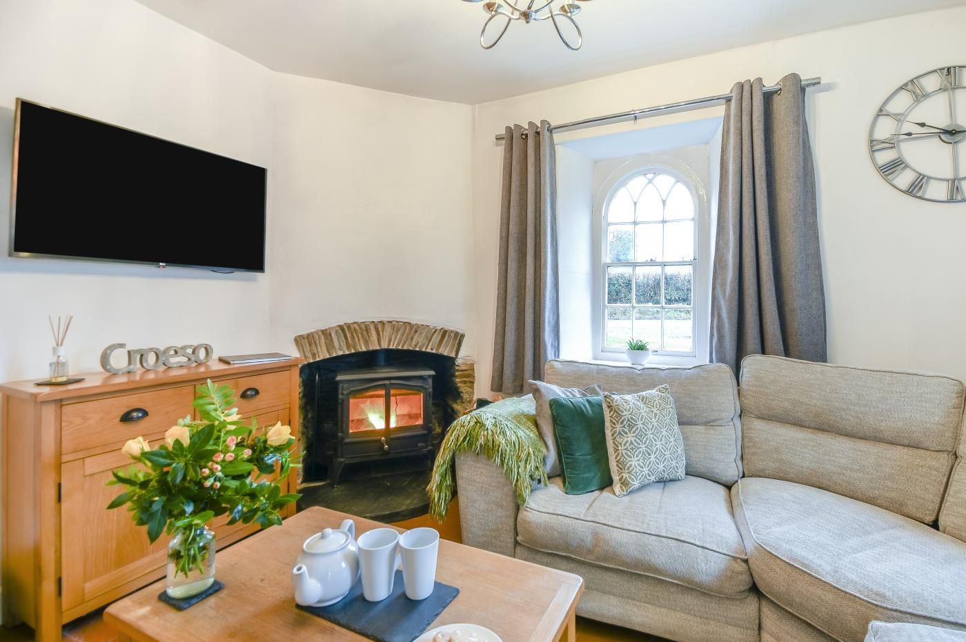 A living room with a fireplace, a sofa, a coffee table with a teapot and cups, and a flat-screen TV mounted on the wall.