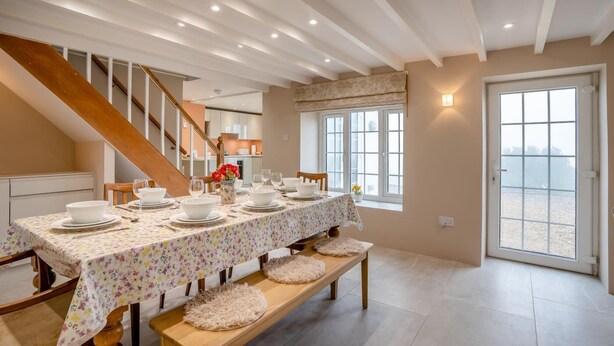 Uma sala de jantar com uma mesa comprida posta para quatro pessoas, uma escada e vista através de uma porta de vidro.