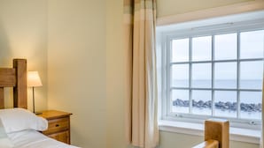 A bedroom with a bed, a nightstand, a lamp, a window with a view of the sea, and a curtain. - 3 Bed in Dunbar (oc-k34653) (Dunbar)