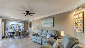 Ein Wohnzimmer mit Sofa, Esstisch, Stühlen, Deckenventilator und einer Wanddekoration im maritimen Stil.