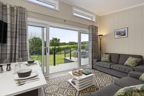 A modern living room with a sofa, a dining table with chairs, and a view of the outdoors through large glass doors.