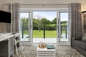 A modern living room with a dining area, a sofa, a TV, and a view of a garden through glass doors.