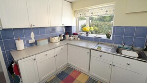 Una cocina con armarios blancos, salpicadero de azulejos azules, ventana con vistas a vegetación, alfombra colorida y un fregadero con escurreplatos.