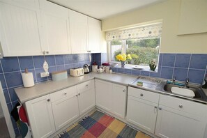 A kitchen with white cabinets, a blue tiled backsplash, a window with a view of greenery, a colorful rug, and a sink with a dish rack. - 2 Bed in Mumbles (oc-hh1203) (The Mumbles)