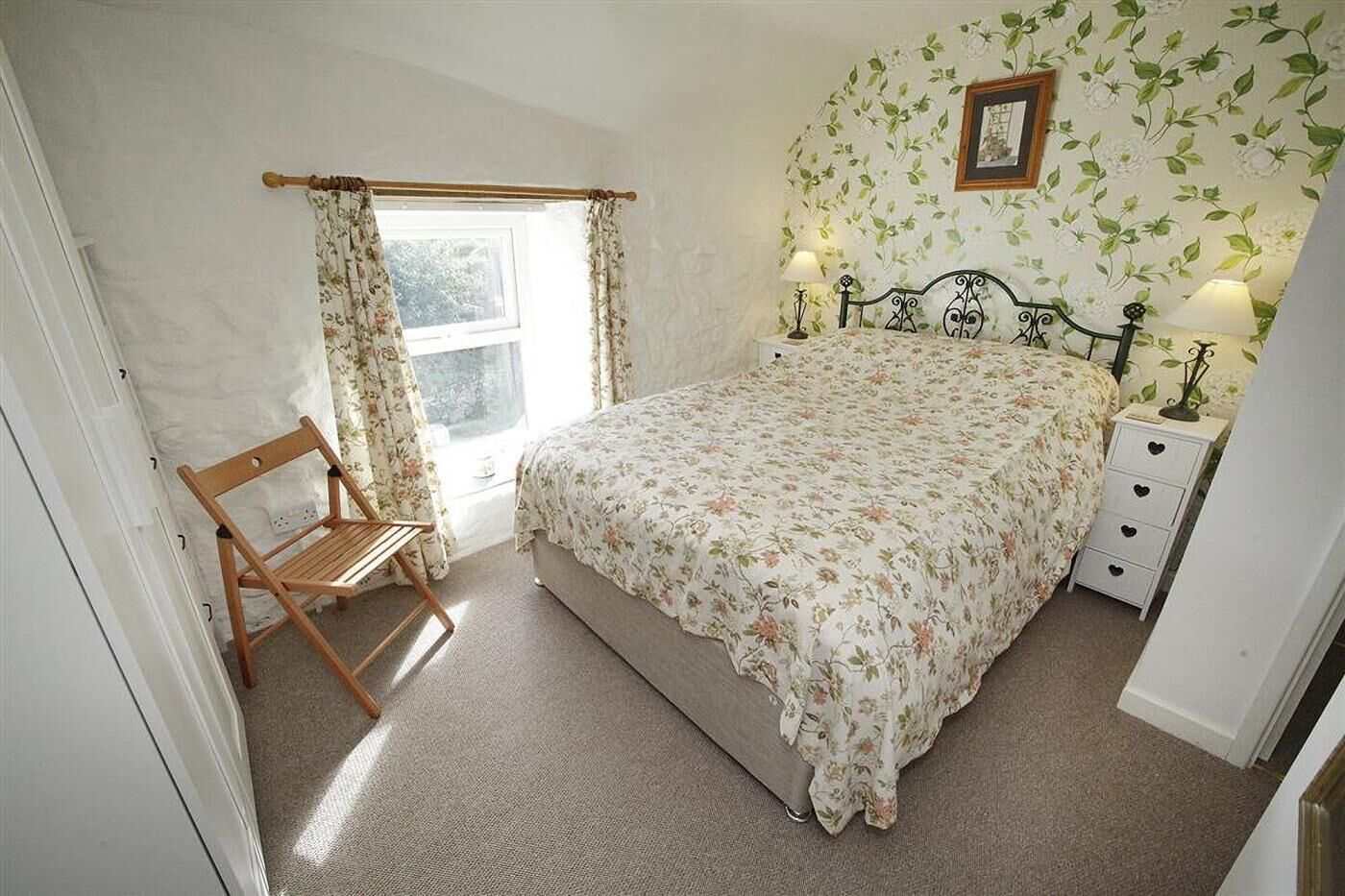 A bedroom with a bed, a chair, a nightstand, a lamp, a window with curtains, and a floral wallpaper.
