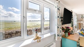 Een kamer met een groot raam met uitzicht op een strand, een kleine tafel met een bloevas en een flatscreen televisie aan de muur.