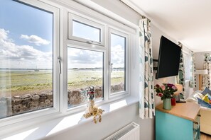 Ein Zimmer mit großem Fenster zum Strand, einem kleinen Tisch mit Blumenvase und einem an der Wand befestigten Flachbildfernseher.