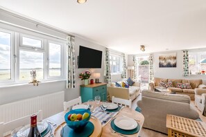 Ein helles Wohnzimmer mit einer Couch, einem Esstisch für zwei Personen und einem an der Wand montierten Fernseher.