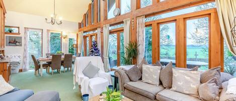 A spacious living room with a sofa, chairs, a dining table, and large windows offering a view of the outdoors.