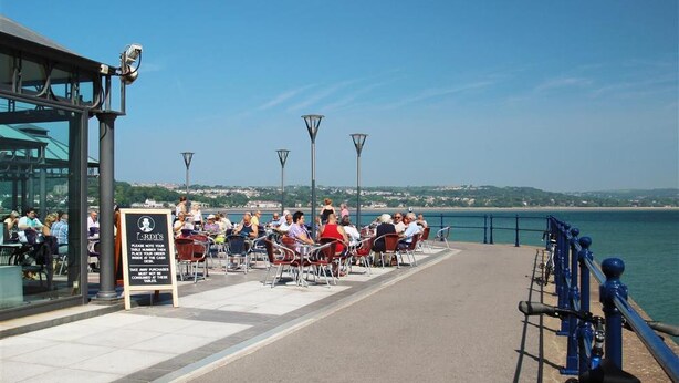 A waterfront dining area with tables and chairs, a signboard, and a view of the sea. - 1 Bed in Mumbles (oc-hh1205) (The Mumbles)