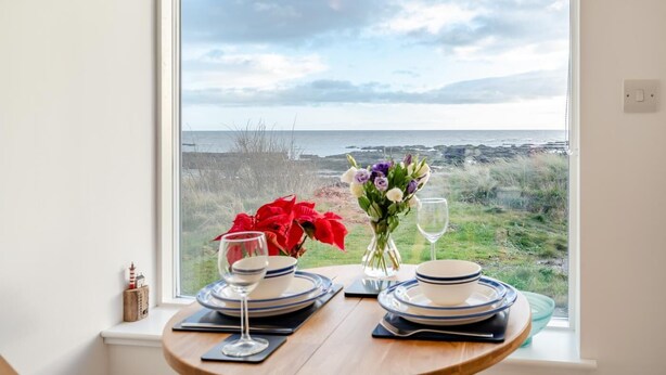 Une table dressée pour deux personnes, avec des assiettes, un verre à vin et un vase de fleurs, donnant sur une vue sur la côte.