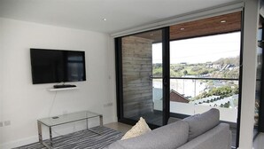 A modern living room with a flat-screen TV, a glass coffee table, a grey sofa, and a large sliding glass door leading to a balcony with a view of a residential area. - 1 Bed in Langland (oc-hh1002) (Newton)