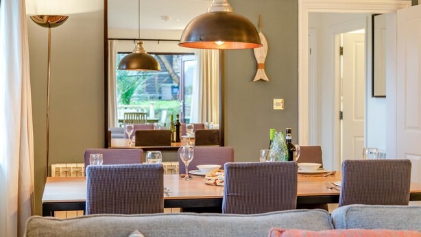 Een eetruimte met een grote tafel, stoelen en een hanglamp. Een bank bevindt zich op de voorgrond en een deur aan de rechterkant.