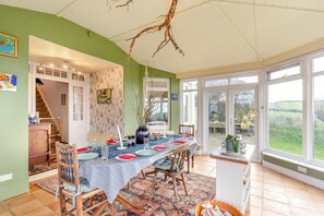Un coin repas avec une longue table dressée pour six personnes, entourée de chaises, d’une nappe à motifs et d’un vase orné de branches.
