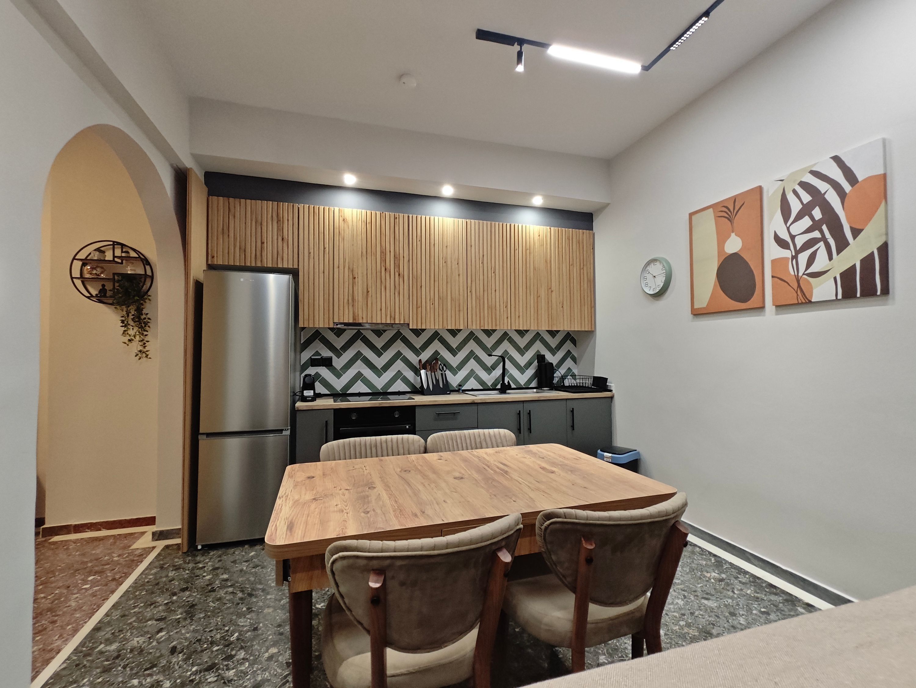 A modern kitchen with a wooden dining table and chairs, a refrigerator, and a herringbone-patterned backsplash.