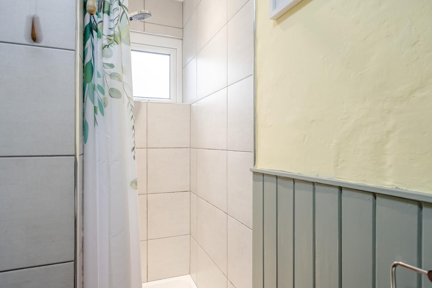 Une salle de bain avec des carreaux blancs, une douche, une fenêtre et une porte dotée d’une poignée.
