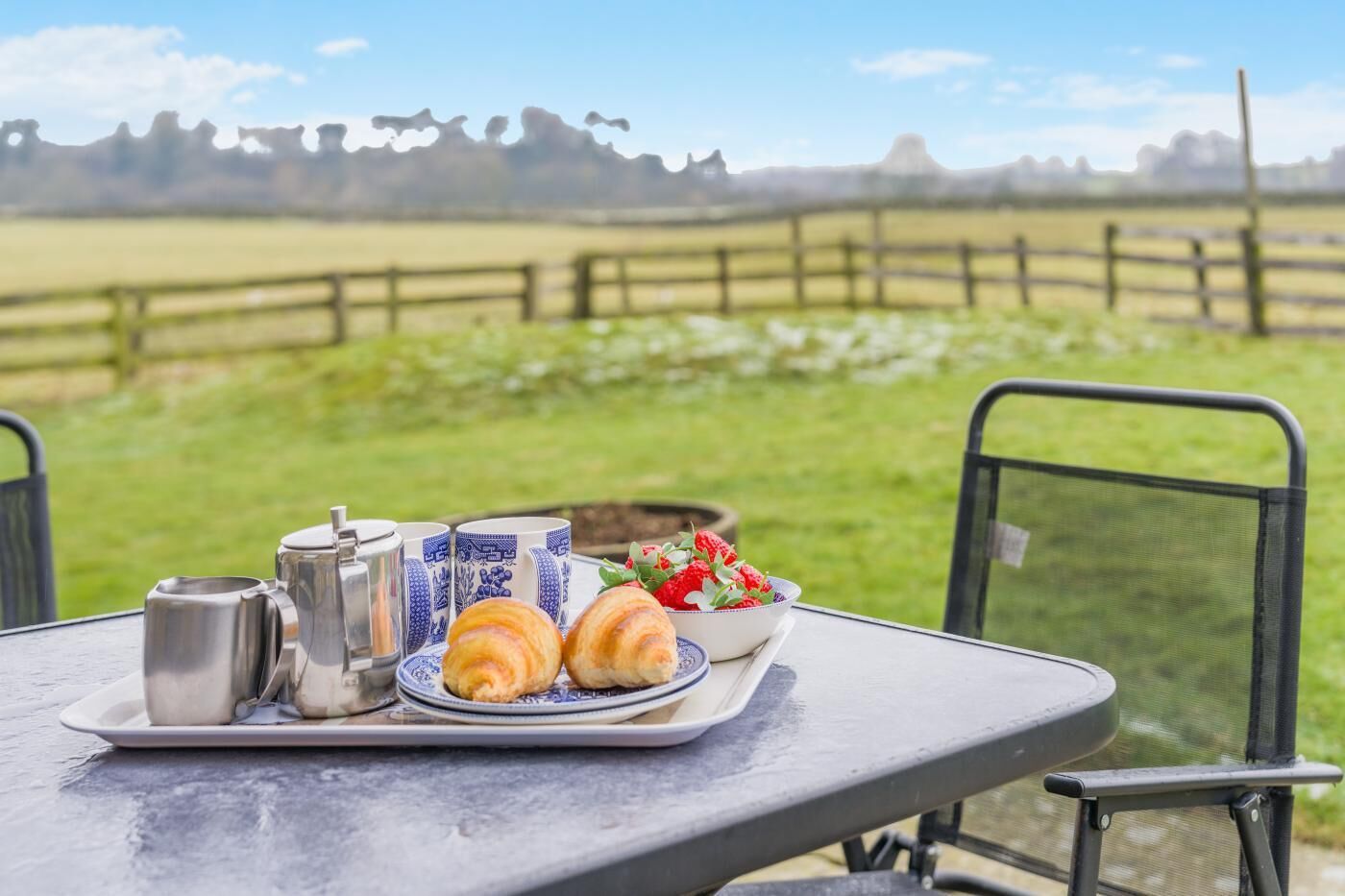 A breakfast tray with croissants, a bowl of strawberries, and two mugs on a table with a view of a grassy field and a wooden fence.
