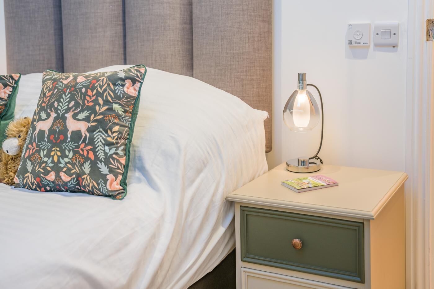 A neatly made bed with floral pillows, a bedside lamp, and a small drawer unit.