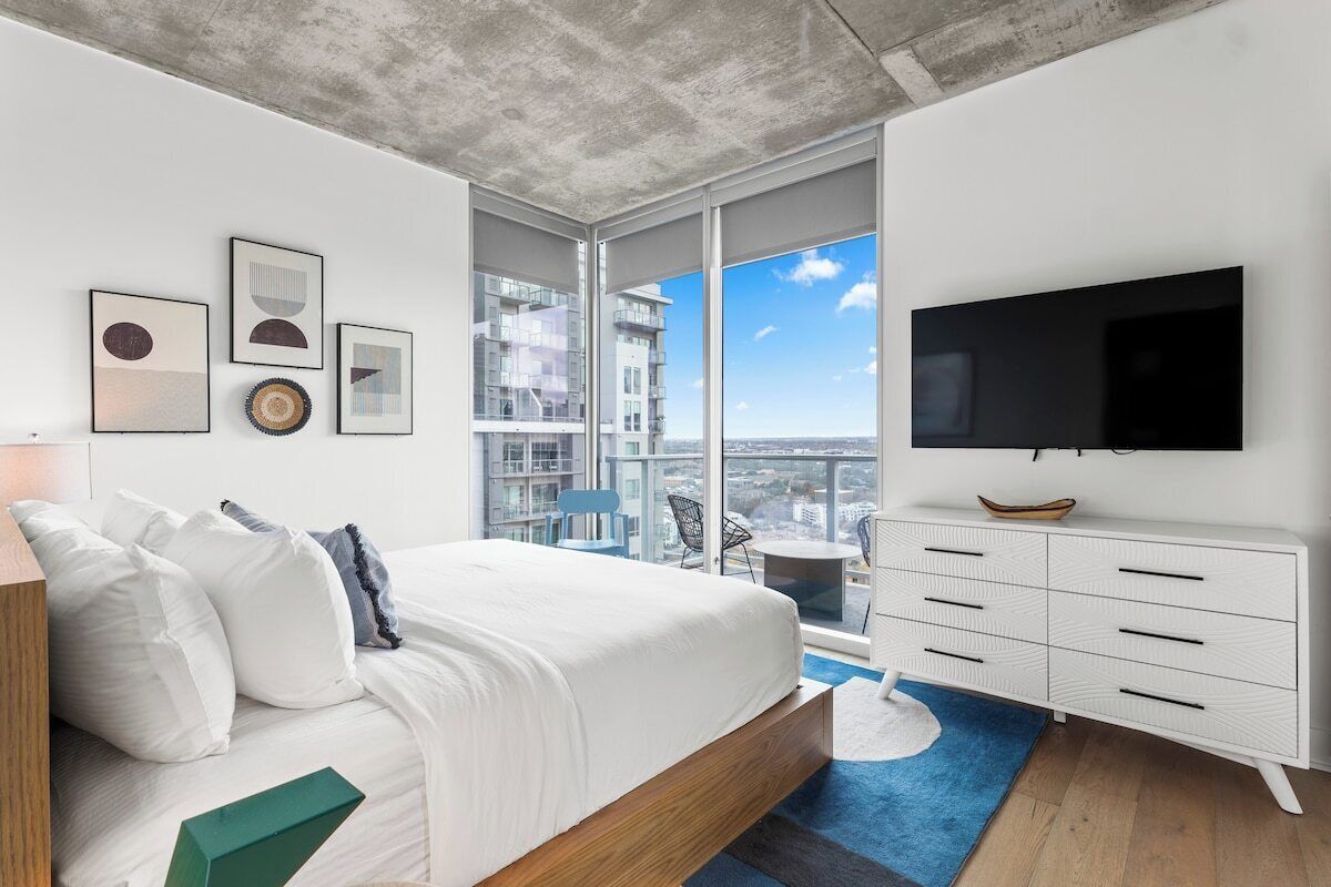 Ein modernes Schlafzimmer mit einem großen Bett, einem Flachbildfernseher, einer weißen Kommode und Blick auf die Stadt.