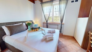A neatly made bed with white linens, a tray with fruits and a mug, a vase with flowers, and a window with curtains. - Lido Apartment by Booking Lake Garda (Manerba del Garda)