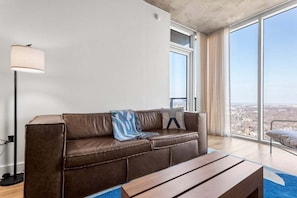 Ein modernes Wohnzimmer mit einem braunen Ledersofa, einem Holztisch, einer Stehlampe und großen Fenstern mit Blick auf die Stadt.