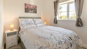 A bedroom with a bed, a nightstand, a lamp, a window with curtains, and a wall-mounted painting.